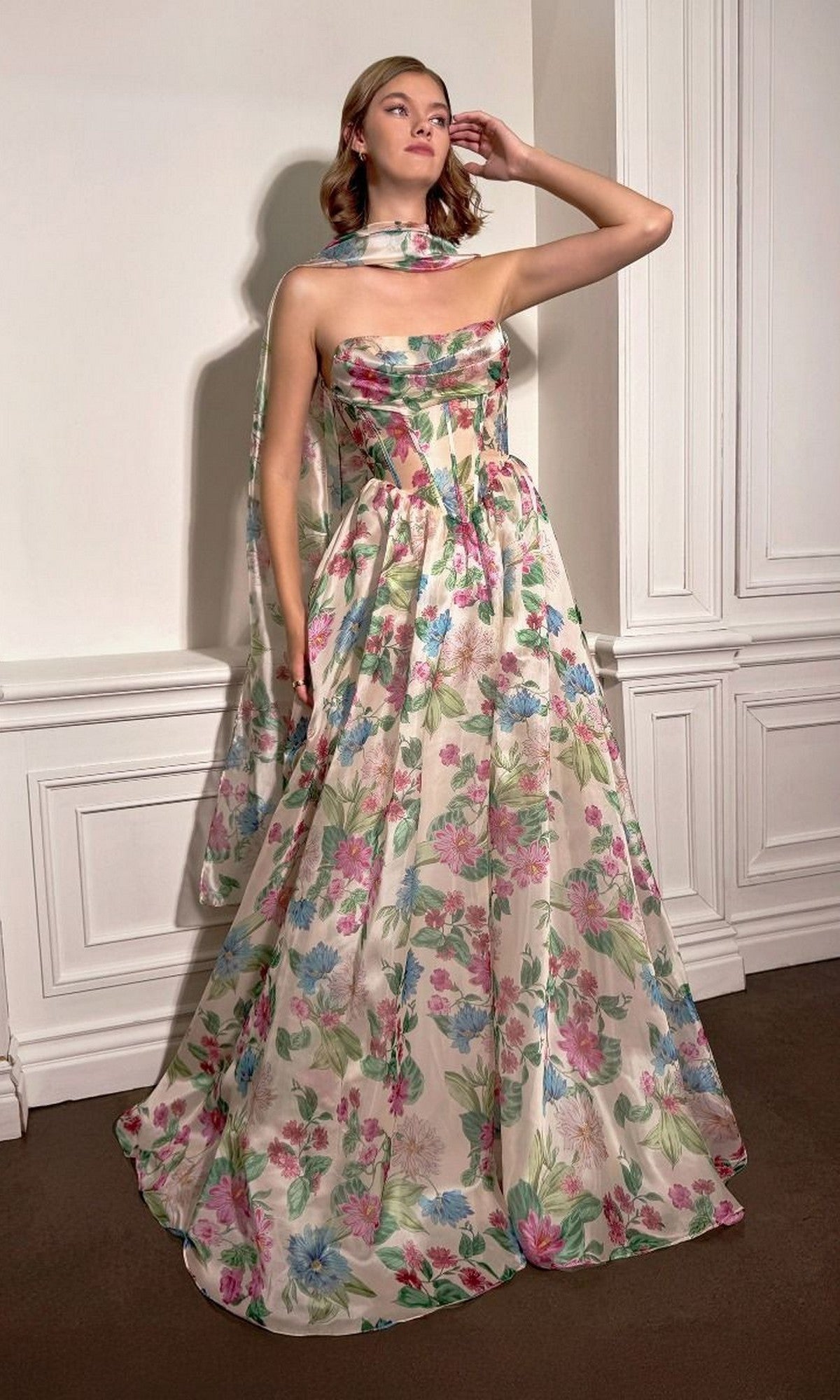 Wearing the Andrea and Leo A1425, a long strapless floral-print prom dress with a corset bodice and matching sheer scarf, the woman poses indoors with one hand gracefully raised near her head.